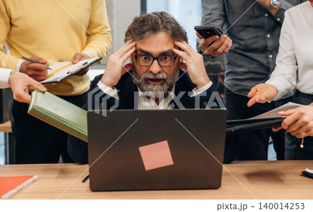 Businessman with glasses is overwhelmed by multiple tasks while working on laptop in a busy office environment, showcasing the challenges of modern workplace dynamics Businessman with glasses is overwhelmed by multiple tasks while working on laptop in a busy office environment, showcasing the challenges of modern workplace dynamics 140014253