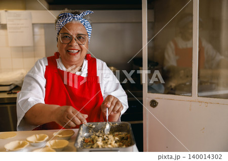 Senior Afro Latina woman smiling while preparing savory pastries in a professional kitchen setting Senior Afro Latina woman smiling while preparing savory pastries in a professional kitchen setting 140014302