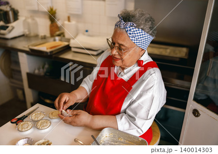 Senior African American woman preparing handmade pastries in a professional commercial kitchen bakery. Senior African American woman preparing handmade pastries in a professional commercial kitchen bakery. 140014303