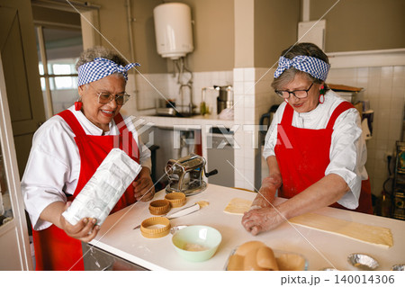 Diverse senior Black and Caucasian women preparing dough in a bright professional kitchen setting Diverse senior Black and Caucasian women preparing dough in a bright professional kitchen setting 140014306