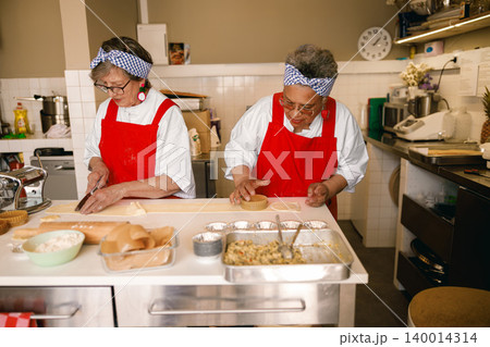 Senior Caucasian and multicultural women preparing pastries in a bright community kitchen workspace Senior Caucasian and multicultural women preparing pastries in a bright community kitchen workspace 140014314