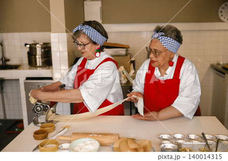 Active senior Caucasian and Black female chefs making fresh pasta in a bright professional kitchen Active senior Caucasian and Black female chefs making fresh pasta in a bright professional kitchen 140014315