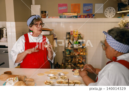 Senior Caucasian and Black women baking small pastries together in a bright professional kitchen. 140014329