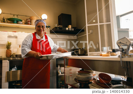 Smiling Hispanic senior woman holding a baking tray in a professional kitchen restaurant setting Smiling Hispanic senior woman holding a baking tray in a professional kitchen restaurant setting 140014330