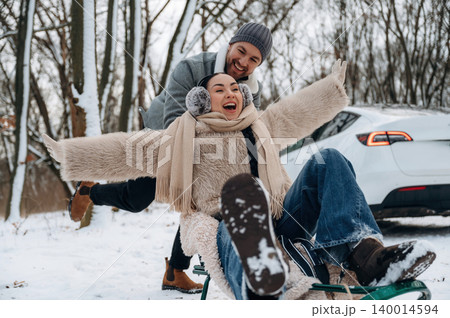 Joyful couple enjoying winter fun on a sled in a snowy landscape, surrounded by trees and a parked vehicle, capturing the essence of outdoor adventure 140014594