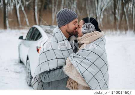 Couple wrapped in cozy blanket sharing a warm embrace in snowy landscape, with a car parked nearby, capturing the essence of winter romance and connection Couple wrapped in cozy blanket sharing a warm embrace in snowy landscape, with a car parked nearby, capturing the essence of winter romance and connection 140014607