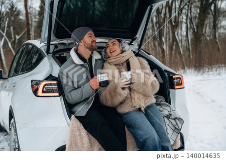 Couple enjoying warm drinks while sitting in the back of a car in a snowy landscape, surrounded by trees, creating a cozy winter atmosphere 140014635
