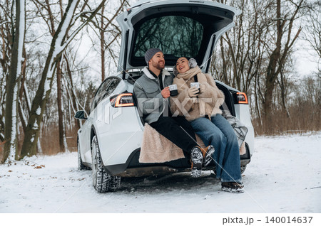 Couple enjoying warm beverages while sitting on the back of a car in a snowy landscape, surrounded by trees, capturing a cozy winter moment Couple enjoying warm beverages while sitting on the back of a car in a snowy landscape, surrounded by trees, capturing a cozy winter moment 140014637