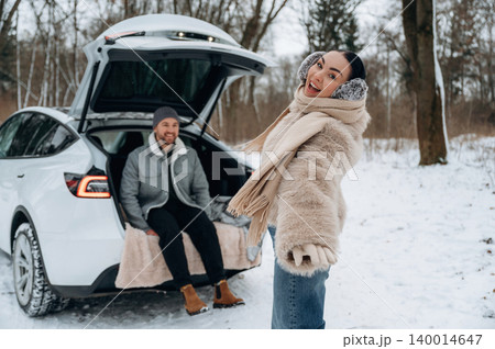 Joyful woman in winter attire poses playfully in snowy landscape near open car trunk, while man relaxes comfortably in background, showcasing winter adventure spirit Joyful woman in winter attire poses playfully in snowy landscape near open car trunk, while man relaxes comfortably in background, showcasing winter adventure spirit 140014647