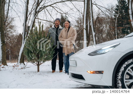 Couple stands beside a Christmas tree in snowy landscape, smiling joyfully near a white electric vehicle, celebrating winter holiday spirit and togetherness Couple stands beside a Christmas tree in snowy landscape, smiling joyfully near a white electric vehicle, celebrating winter holiday spirit and togetherness 140014764