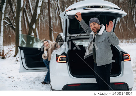 Happy couple enjoying winter day outdoors, standing by open trunk of electric vehicle in snowy forest, showcasing joyful moments and adventure Happy couple enjoying winter day outdoors, standing by open trunk of electric vehicle in snowy forest, showcasing joyful moments and adventure 140014804