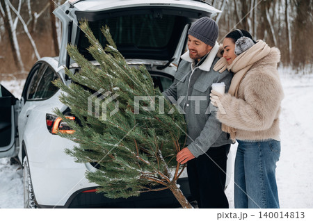 Couple enjoying winter holiday tradition, loading freshly cut Christmas tree into car trunk, surrounded by snow-covered landscape and trees, capturing festive spirit Couple enjoying winter holiday tradition, loading freshly cut Christmas tree into car trunk, surrounded by snow-covered landscape and trees, capturing festive spirit 140014813