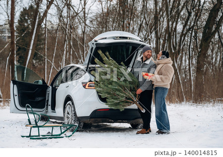 Couple enjoying winter holiday season, unloading Christmas tree from car in snowy landscape, surrounded by nature and festive atmosphere with sled nearby 140014815