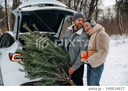 Couple enjoying winter holiday season while loading Christmas tree and gift into car trunk, surrounded by snowy landscape and trees, capturing festive spirit Couple enjoying winter holiday season while loading Christmas tree and gift into car trunk, surrounded by snowy landscape and trees, capturing festive spirit 140014817