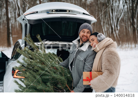 Couple joyfully loading a Christmas tree and gift into a car trunk, surrounded by a snowy winter landscape, capturing the festive holiday spirit and togetherness 140014818