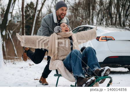 Joyful couple enjoying winter fun on sled in snowy landscape, with car parked nearby, capturing the essence of outdoor adventure and seasonal excitement Joyful couple enjoying winter fun on sled in snowy landscape, with car parked nearby, capturing the essence of outdoor adventure and seasonal excitement 140014856