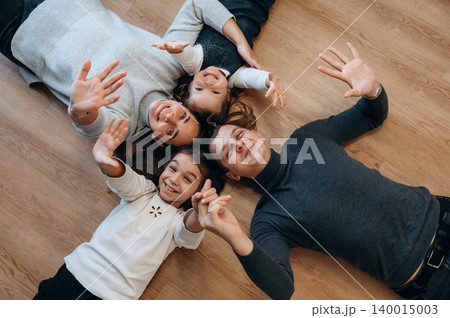 Family enjoying quality time together, lying on wooden floor, smiling and waving hands, capturing joyful moments of togetherness and happiness Family enjoying quality time together, lying on wooden floor, smiling and waving hands, capturing joyful moments of togetherness and happiness 140015003