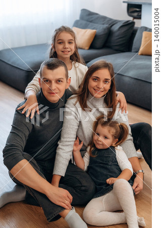Family portrait featuring parents and two daughters sitting together on a cozy living room floor, showcasing warmth and togetherness in a modern home environment Family portrait featuring parents and two daughters sitting together on a cozy living room floor, showcasing warmth and togetherness in a modern home environment 140015004