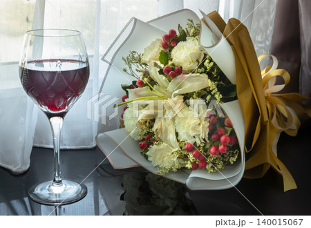 an elegant stemmed glass filled halfway with deep red wine stands on a dark, glossy table beside a sheer white curtain. a beautifully wrapped bouquet rests horizontally next to the glass. an elegant stemmed glass filled halfway with deep red wine stands on a dark, glossy table beside a sheer white curtain. a beautifully wrapped bouquet rests horizontally next to the glass. 140015067