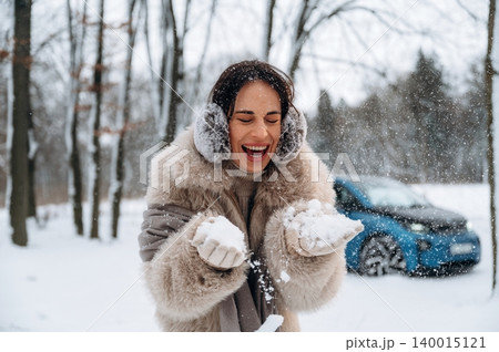 Woman with dark hair, wearing a cozy fur coat and earmuffs, joyfully playing with snow in a winter landscape, surrounded by trees and a parked vehicle in the background Woman with dark hair, wearing a cozy fur coat and earmuffs, joyfully playing with snow in a winter landscape, surrounded by trees and a parked vehicle in the background 140015121