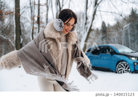 Young woman in winter attire joyfully dancing in snow-covered forest, showcasing warm clothing and playful spirit, with electric car parked in the background Young woman in winter attire joyfully dancing in snow-covered forest, showcasing warm clothing and playful spirit, with electric car parked in the background 140015123