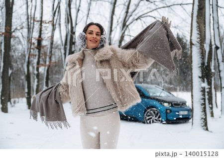 Woman with curly hair, wearing a stylish fur coat and earmuffs, joyfully poses in a snowy forest, showcasing winter fashion and a blue electric car in the background Woman with curly hair, wearing a stylish fur coat and earmuffs, joyfully poses in a snowy forest, showcasing winter fashion and a blue electric car in the background 140015128