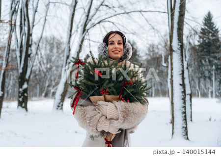 Smiling woman with earmuffs holding a festive bouquet of evergreen branches adorned with red ribbons, standing in a snowy winter landscape surrounded by trees, celebrating the holiday season Smiling woman with earmuffs holding a festive bouquet of evergreen branches adorned with red ribbons, standing in a snowy winter landscape surrounded by trees, celebrating the holiday season 140015144