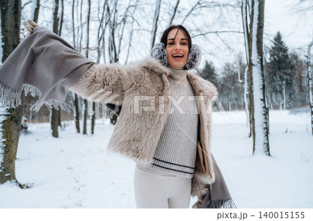 Young woman with dark hair, wearing a cozy fur coat and earmuffs, joyfully poses in a snowy forest, showcasing winter fashion and a cheerful atmosphere in nature Young woman with dark hair, wearing a cozy fur coat and earmuffs, joyfully poses in a snowy forest, showcasing winter fashion and a cheerful atmosphere in nature 140015155