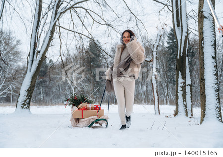 Woman in winter attire joyfully pulls a sled filled with holiday gifts through a snowy landscape, surrounded by trees, capturing the essence of festive cheer and winter wonder Woman in winter attire joyfully pulls a sled filled with holiday gifts through a snowy landscape, surrounded by trees, capturing the essence of festive cheer and winter wonder 140015165