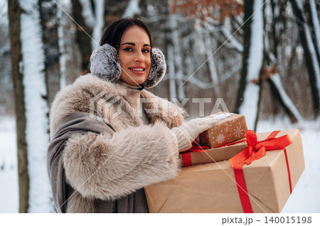 Woman in winter attire, wearing earmuffs, carries beautifully wrapped gifts in a snowy forest, showcasing festive spirit and holiday joy in a serene outdoor setting Woman in winter attire, wearing earmuffs, carries beautifully wrapped gifts in a snowy forest, showcasing festive spirit and holiday joy in a serene outdoor setting 140015198