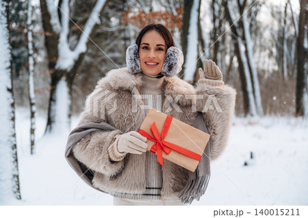 Woman in winter attire, holding a gift wrapped with a red ribbon, smiles cheerfully in a snowy forest, embodying the spirit of holiday celebrations and joy 140015211