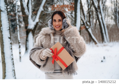 Woman with dark hair wearing a cozy fur coat and earmuffs, holding a gift box with a red ribbon, standing in a snowy forest, celebrating winter holidays with joy and warmth 140015212