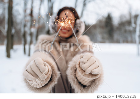 Woman in winter attire holding sparklers in snowy landscape, showcasing festive spirit and joy, surrounded by trees, celebrating the holiday season with warmth and light Woman in winter attire holding sparklers in snowy landscape, showcasing festive spirit and joy, surrounded by trees, celebrating the holiday season with warmth and light 140015219