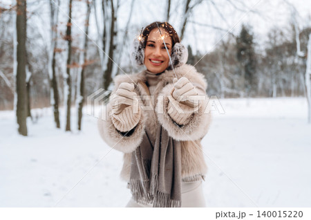 Smiling woman in winter attire holding sparklers in a snowy forest, surrounded by trees, celebrating the holiday season with joy and warmth in a festive atmosphere Smiling woman in winter attire holding sparklers in a snowy forest, surrounded by trees, celebrating the holiday season with joy and warmth in a festive atmosphere 140015220