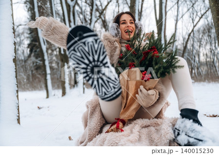 Young woman with brown hair, wearing winter clothing, joyfully poses in snowy forest, holding festive greenery and smiling, capturing the essence of winter celebration and joy Young woman with brown hair, wearing winter clothing, joyfully poses in snowy forest, holding festive greenery and smiling, capturing the essence of winter celebration and joy 140015230