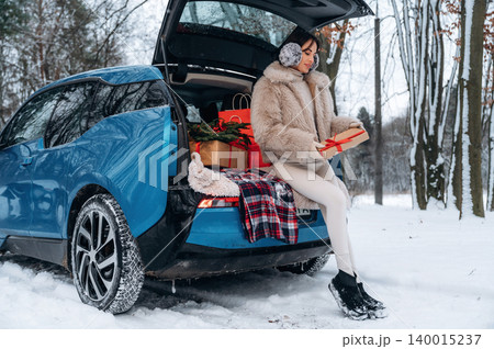 Woman in winter attire sitting on car trunk, holding gift box surrounded by holiday decorations, snow-covered landscape creating a festive atmosphere with copy space Woman in winter attire sitting on car trunk, holding gift box surrounded by holiday decorations, snow-covered landscape creating a festive atmosphere with copy space 140015237