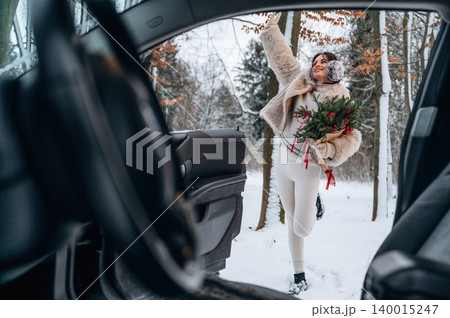 Woman in winter attire, holding festive greenery, stepping out of a car in a snowy landscape, capturing the essence of holiday spirit and outdoor adventure Woman in winter attire, holding festive greenery, stepping out of a car in a snowy landscape, capturing the essence of holiday spirit and outdoor adventure 140015247