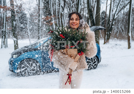Woman with dark hair holding festive greenery in snowy landscape, wearing cozy winter attire, standing beside a blue electric vehicle, celebrating holiday spirit in nature Woman with dark hair holding festive greenery in snowy landscape, wearing cozy winter attire, standing beside a blue electric vehicle, celebrating holiday spirit in nature 140015266