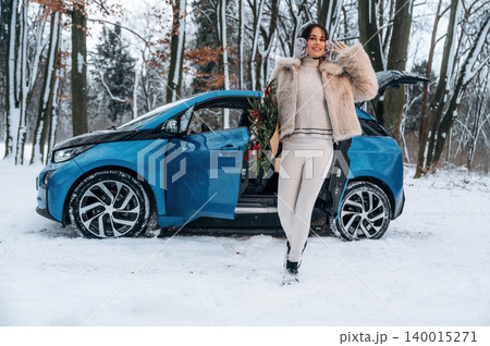 Stylish woman in winter attire poses beside a blue electric car in a snowy forest, showcasing eco-friendly transportation and seasonal beauty with holiday decorations 140015271