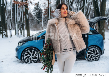 Woman in stylish winter attire, holding festive greenery, stands beside a blue electric car in a snowy forest, showcasing seasonal joy and modern transportation Woman in stylish winter attire, holding festive greenery, stands beside a blue electric car in a snowy forest, showcasing seasonal joy and modern transportation 140015272