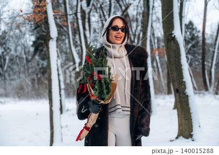 Stylish woman in winter attire, holding a festive bouquet, smiles joyfully in a snowy forest, surrounded by tall trees and a serene winter landscape, celebrating the holiday season Stylish woman in winter attire, holding a festive bouquet, smiles joyfully in a snowy forest, surrounded by tall trees and a serene winter landscape, celebrating the holiday season 140015288