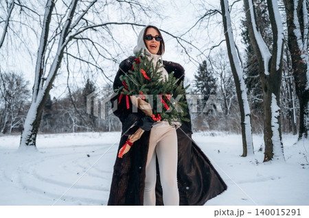 Stylish woman in a luxurious fur coat, holding a festive wreath adorned with red ribbons, joyfully walking through a snowy landscape, celebrating the winter holiday season with elegance and cheer Stylish woman in a luxurious fur coat, holding a festive wreath adorned with red ribbons, joyfully walking through a snowy landscape, celebrating the winter holiday season with elegance and cheer 140015291