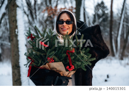 Woman in sunglasses, wearing a stylish coat, holds a festive bouquet of evergreen branches with red berries, surrounded by a snowy winter landscape, celebrating the holiday season 140015301