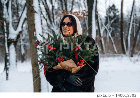 Woman in winter attire holding a festive bouquet of evergreen branches with red berries, surrounded by a snowy forest, celebrating the holiday season with joy and warmth 140015302
