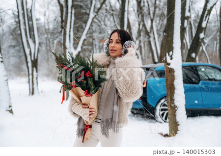 Woman with dark hair, wearing cozy winter attire, holding festive greenery bouquet in snowy forest, with blue car parked nearby, celebrating holiday spirit and winter joy 140015303