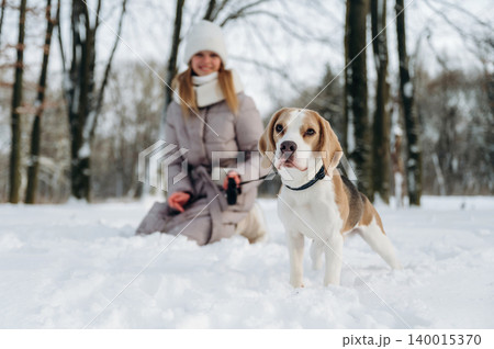 Woman in winter coat kneeling in snow with a beagle dog on a leash surrounded by trees in a snowy park during a cold day with soft natural lighting Woman in winter coat kneeling in snow with a beagle dog on a leash surrounded by trees in a snowy park during a cold day with soft natural lighting 140015370
