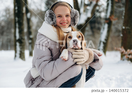 Young girl with long blonde hair wearing earmuffs and a winter coat is holding a beagle dog in a snowy park surrounded by trees during a cold day 140015372