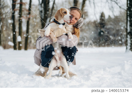 Young woman with long hair wearing earmuffs is holding a beagle dog in a snowy park, surrounded by trees and falling snowflakes during winter season Young woman with long hair wearing earmuffs is holding a beagle dog in a snowy park, surrounded by trees and falling snowflakes during winter season 140015374