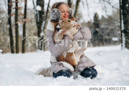 Young girl wearing earmuffs is sitting in the snow while holding a beagle dog, surrounded by trees in a winter landscape during a joyful outdoor moment Young girl wearing earmuffs is sitting in the snow while holding a beagle dog, surrounded by trees in a winter landscape during a joyful outdoor moment 140015378