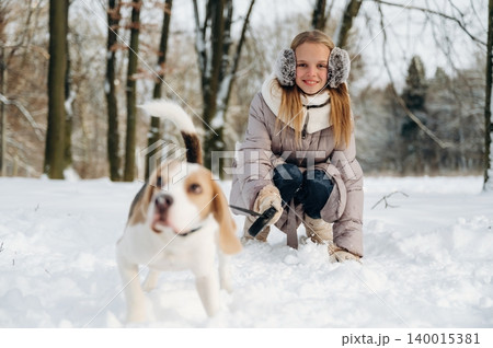 Young girl with long blonde hair wearing earmuffs is playing with a beagle dog in a snowy forest, trees visible in the background during winter outdoor activity 140015381
